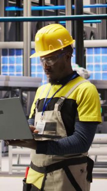 Güneş panelleri fabrikasındaki gülümseyen teknisyenin dikey video portresi robot montaj hatlarını denetliyor ve kalite kontrolü sağlıyor. Endüstriyel fabrikadaki Afrikalı Amerikalı adam otomatik taşıyıcıyı izliyor