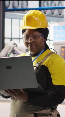 Dikey video gülümseyen akıllı fabrika mühendisi dizüstü bilgisayarda yazıyor, üretim hatlarının izlenmesini denetliyor. Sektör 4.0 fabrikasındaki neşeli kadın makinelerin en uygun verimlilikte çalışmasını sağlıyor.