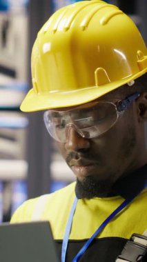 Akıllı fabrikada çalışan dikey video uzmanı güvenilirliğini sağlamak için otomatik sistemleri test ediyor. Endüstri 4.0 'daki Afrikalı Amerikalı adam not defterini kullanarak darboğazları tespit ediyor ve optimize ediyor.