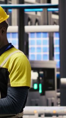 Akıllı fabrikadaki dikey video teknisyeni sistem performans ölçümlerine bakmak için defter kullanıyor. IoT sensörleri aracılığıyla enerji üretimini takip etmek için dizüstü bilgisayar kullanan 4.0 sanayiindeki Afrikalı Amerikalı adam.