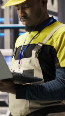 Dikey video Akıllı güneş panelleri fabrika teknisyeni dizüstü bilgisayarını taşıyıcı bantları denetlemek için kullanıyor. Endüstrideki Afrikalı Amerikalı adam 4.0 fabrika makinelerinin en uygun verimlilikte çalıştığından emin oluyor.