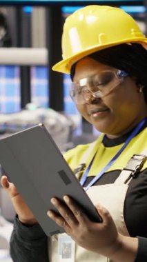 Güneş panellerinde çalışan dikey video kadın makine sistemlerini denetlemek için tablet kullanıyor. Afrikalı Amerikalı fotovoltaik fabrikası çalışanı işlemleri sürdürmek için yazılımı kullanıyor, kamera B