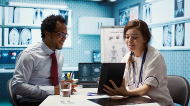 Tıbbi uzman kabinedeki test sonuçlarını açıklıyor. İyileşme sürecindeki adama yardım etmek için röntgen görüntülerini inceliyor. Sağlık ekibi muayene sırasında tarama yorumu yapıyor. Kamera B.