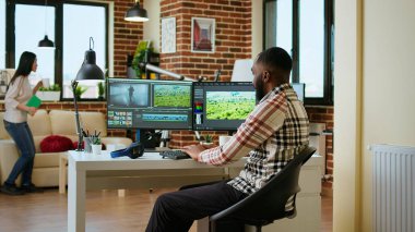 Erkek video editörü, oturma odasındaki modern ev ofisinden çifte monitör kurulumu ve düzenleme yazılımı ile birden fazla iş yapıyor. Medya üretimi için üretim sonrası görevler tamamlanıyor. Kamera A.