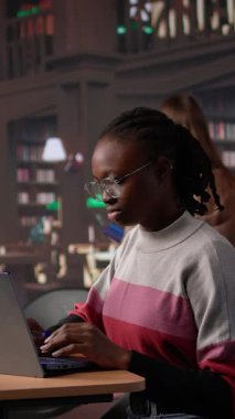 Dikey Video Afrikalı Amerikalı öğrenci kütüphanede akademik araştırma koleksiyonuna danışmanlık yapıyor, üniversite diplomasını almak için yaklaşan sınavlara çalışıyor. Kız burslu çalışıyor, eğitim görüyor. Kamera.