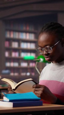 Dikey Video Siyah kız, kampüs kütüphanesinde sınav hazırlıklarına odaklanıyor, laptopuna barışçıl bir çalışma ortamındaki referans kitaplarından notlar alıyor. Kız genel veritabanı materyallerini kullanıyor