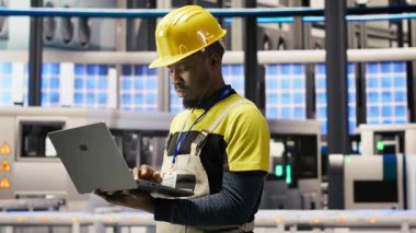 Akıllı fabrikadaki teknisyen sistem performans ölçümlerine bakmak için defter kullanıyor. IoT sensörleri ve kamera B aracılığıyla enerji üretimini takip etmek için dizüstü bilgisayar kullanan 4.0 sanayiindeki Afrikalı Amerikalı adam.