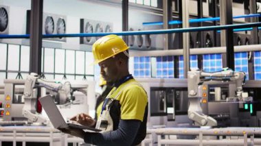 Akıllı fabrikadaki teknisyen sistem performans ölçümlerine bakmak için defter kullanıyor. IoT sensörleri ve kamera A aracılığıyla enerji üretimini takip etmek için dizüstü bilgisayar kullanan 4.0 sanayiindeki Afrikalı Amerikalı adam.