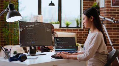 Uzman bilişim uzmanı uzaktan çalışırken rahat bir ofisteki çift monitörlere kod yazar. Asyalı kadın kodlamada ve internet güvenliğinde verimlilik için programlama dili kullanır. Kamera B.