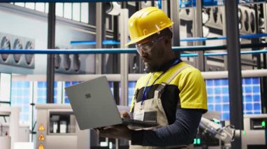 Akıllı fabrikada profesyonelce çalışmak, güvenilirliği sağlamak için otomatik sistemleri test etmek. Endüstri 4.0 'daki Afrikalı Amerikalı adam not defterini kullanarak darboğazları tespit edip optimize ediyor. Kamera A