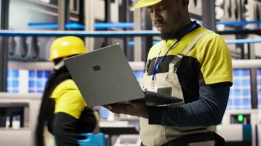 Akıllı güneş panelleri fabrika teknisyeni, dizüstü bilgisayarını taşıma bantlarını denetlemek için kullanıyor. Endüstrideki Afrikalı Amerikalı adam 4.0 fabrika makinelerinin en uygun verimlilikte çalıştığını garanti ediyor. Not defteri, kamera B kullanarak.