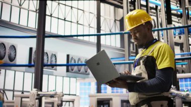 Akıllı güneş panelleri fabrika teknisyeni, dizüstü bilgisayarını taşıma bantlarını denetlemek için kullanıyor. Endüstrideki Afrikalı Amerikalı adam 4.0 fabrika makinelerinin en uygun verimlilikte çalıştığını garanti ediyor. Not defteri, kamera A kullanarak.