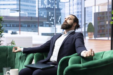 Modern bir ofiste iş adamı meditasyon müziğiyle gevşer, yoğun programında uyumu ve sakinliği bulur. Rahat duruşu olan CEO, kendine bakmanın önemini vurguluyor.