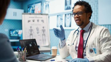 Afrikalı Amerikalı doktor lastik eldivenlerini takıyor ve kabinede kalp şeklini gösteriyor. Hasta bakımını ve profesyonel sağlık hizmetlerini vurguluyor. Kontroldeki doktor. Kamera B.