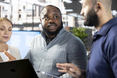 İşçilerle birlikte dizüstü bilgisayar ekranına bakan yönetici, onlara iş görüşlerini bulmak ve projeyi bitirmek için gerekli anahtar verileri araştırırken gösteriyor. Not defterindeki verileri analiz eden yönetici yöneticileri