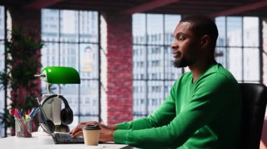 Çatı katındaki bilgisayarında yazı yazan Afro-Amerikan bir yetişkin. Çevrimiçi serbest çalışma ve verimli bir şekilde birçok iş yapıyor. Rahat bir ofiste uzaktan kumandanın esnekliği. Kamera A.