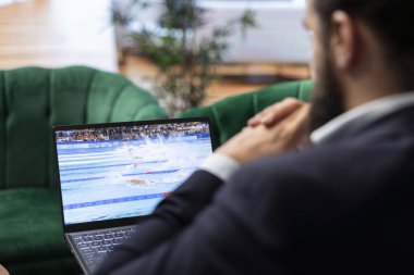Bükreş, Romanya - 30 Temmuz 2024 - Yönetmen ulusal atlet ve şampiyonu desteklemek amacıyla Olimpiyatların yüzme duruşmasını izliyor. Genç CEO canlı yayını dizüstü bilgisayarda kullanıyor..