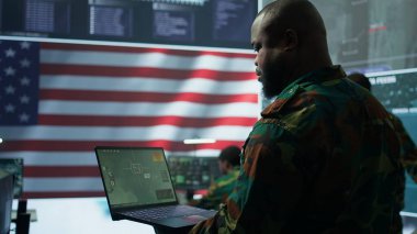 Amerikan askeri üniformalı istihbarat memurları bir komuta merkezinde bilgisayar üzerinde çalışıyor, gözlem ve veri analizini yönetiyorlar. ABD hükümeti için küresel operasyonları destekleyin. Kamera B.