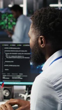 Dikey video sunucusu çiftliği bilgisayar bilimcisi yapay zeka veri analizi grafiklerini yorumluyor. Veri merkezinde tahmini sistem ölçümleri gösteren yapay zeka programı kullanan Afrikalı Amerikalı bir adam.