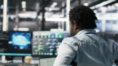 Paraplejik sunucu odası programcısı yapay zeka veri analizi grafiklerini yorumluyor. Tekerlekli sandalyedeki adam, veri merkezinde öngörü sistemi ölçümleri gösteren yapay zeka programı kullanıyor. Kamera B