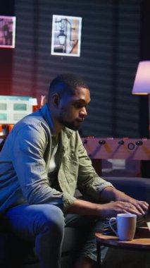 Dikey Afrikalı Amerikalı genç yetişkin bilgisayarla uzaktan kumandalı çatı katı ışıklarının altında çalışıyor, rahat dairesinde bir kanepede oturuyor. Şık bir evde rahat bir insan modern üretkenliği ön plana çıkarıyor