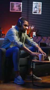 Dikey Afrikalı Amerikalı adam dizüstü bilgisayarda çalışıp neon ışıkların altında başarıyı kutluyor, rahat bir dairede kanepede oturuyor. Rahatlamış insan müzik dinler, görevleri başarıyla tamamlar.