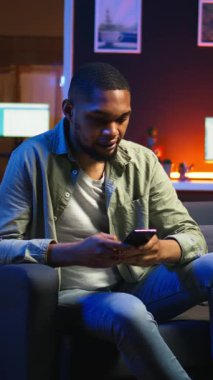Dikey Video Afrikalı Amerikalı bir insan koltukta akıllı telefon uygulaması üzerinden mesaj atıyor, evde dinlenmeden önce görevlerini tamamlıyor. Genç yetişkin cep telefonunu kaydırıyor, mesajlarla sohbet ediyor. Kamera A.