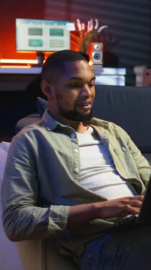 Dikey Video Yetişkinleri dizüstü bilgisayarında yazan adam şık çatı katında kanepeye uzanmış, serbest ve çevrimiçi işlere girişiyor. Siyahi kişi uzak işin esnekliğini benimsiyor ve çoklu görev yapıyor