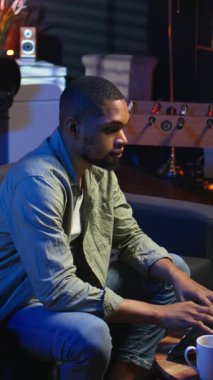 Dikey video odaklı Afrikalı Amerikalı adam şık bir oturma odasında dizüstü bilgisayarıyla telgraf çekiyor. Modern bir çalışma alanında konfor ve verimliliği dengeleyen uzaktan kumandalı bir işçi. Rahat çatı katı stüdyosu