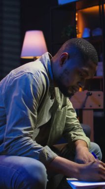 Dikey Video Siyah Serbest Çalışan, ödevlerini çözmek, araştırma yapmak ve makaleleri çevrimiçi okumak için ders kitabı üzerine notlar alıyor. Uzaktan kumandalı işten ve gevşek programdan hoşlanan genç yetişkinler.