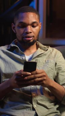 Dikey Video Genç Siyah Adam sosyal medya mesajlaşma uygulamaları üzerinden arkadaşlarına mesaj atıyor, akıllı telefonunu kullanarak internete kayıyor. Odaklanmış biri telefonuyla rahatlıyor, oturma odasında oturuyor.