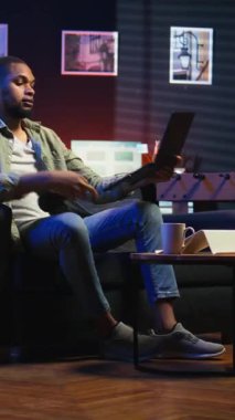 Dikey Video Siyah telgraf işçisi Webinar tartışmasını dinlemek için kulaklık takıyor, yeteneklerini geliştirmek için dizüstü bilgisayar dersine katılıyor. Genç adam oturma odasında buluşmaktan zevk alıyor.