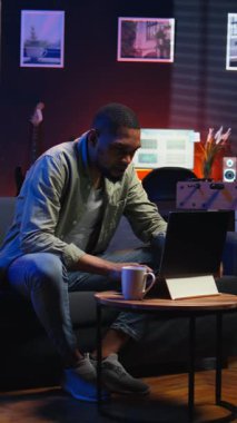 Dikey videolu yetişkin adam dizüstü bilgisayarda yazıyor şık bir dairede koltukta yatıyor, serbest çalışmalar yapıyor ve internetten çalışıyor. Siyahi kişi birden fazla görevi verimli bir şekilde yerine getiriyor ve uzak işin esnekliğini kucaklıyor