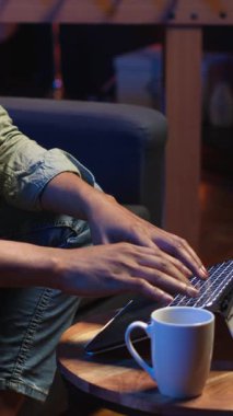 Dikey Afro-Amerikan genç yetişkin bir bilgisayar üzerinde çalışıyor, çatı katı ışıklarının altında, rahat dairesinde bir kanepede oturuyor. Şık bir evde rahat bir insan modern olanları ön plana çıkarıyor.