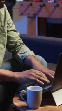 Dikey video odaklı Afrikalı Amerikalı adam şık bir oturma odasında dizüstü bilgisayarıyla telgraf çekiyor. Modern bir çalışma alanında konfor ve verimliliği dengeleyen uzaktan kumandalı bir işçi. Rahat çatı katı stüdyosu