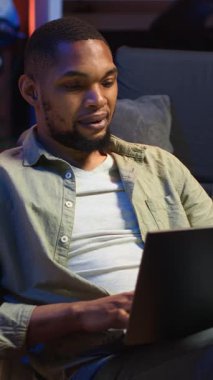 Dikey Video Yetişkinleri dizüstü bilgisayarında yazan adam şık çatı katında kanepeye uzanmış, serbest ve çevrimiçi işlere girişiyor. Siyahi kişi uzak işin esnekliğini benimsiyor ve çoklu görev yapıyor