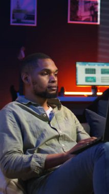 Dikey Afrikalı Afrikalı Amerikalı adam dizüstü bilgisayarıyla uzaktan kumandalı olarak çalışıyor, fasulye torbasının üzerine uzanmış vazifelerini tamamlamaya odaklanıyor. Üretkenlik ve rahatlama ile telgrafı harmanlamak. Kamera A.