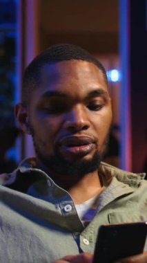 Dikey Video Afrikalı Amerikalı genç adam telefonuyla kanepede mola veriyor, arkadaşlarına sosyal medyada mesaj atıyor ve eğleniyor. Modern oturma odası uzak yaşam tarzında eğlence sunar.