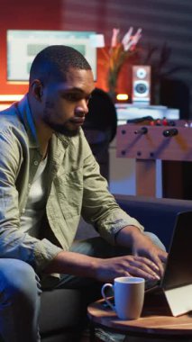 Dikey Afrikalı Amerikalı genç yetişkin bilgisayarla uzaktan kumandalı çatı katı ışıklarının altında çalışıyor, rahat dairesinde bir kanepede oturuyor. Şık bir evde rahat bir insan modern üretkenliği ön plana çıkarıyor