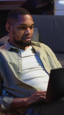 Dikey Video Genç siyah kişi dizüstü bilgisayarında uzaktan görüntülü arama yapıyor evde oturuyor, görevleri çözmek için online konferans webinarına katılıyor. Oturma odasında telekonferans. Kamera.