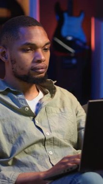 Dikey Afrikalı Afrikalı Amerikalı adam dizüstü bilgisayarıyla uzaktan kumandalı olarak çalışıyor, fasulye torbasının üzerine uzanmış vazifelerini tamamlamaya odaklanıyor. Üretkenlik ve rahatlama ile telgrafı harmanlamak. Kamera B.