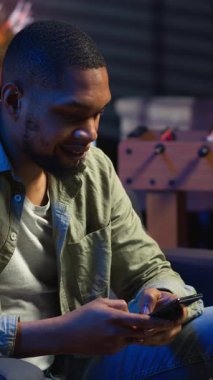 Dikey Video Afrikalı Amerikalı bir insan koltukta akıllı telefon uygulaması üzerinden mesaj atıyor, evde dinlenmeden önce görevlerini tamamlıyor. Genç yetişkin cep telefonunu kaydırıyor, mesajlarla sohbet ediyor. Kamera B.