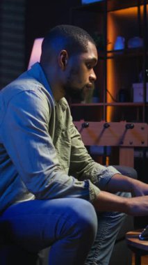 Dikey Afro-Amerikan genç yetişkin bir bilgisayar üzerinde çalışıyor, çatı katı ışıklarının altında, rahat dairesinde bir kanepede oturuyor. Şık bir evde rahat bir insan modern olanları ön plana çıkarıyor.