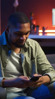 Dikey Video Afrikalı Amerikalı bir insan koltukta akıllı telefon uygulaması üzerinden mesaj atıyor, evde dinlenmeden önce görevlerini tamamlıyor. Genç yetişkin cep telefonunu kaydırıyor, mesajlarla sohbet ediyor. Kamera B.