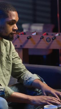 Dikey Video Genç Zenci, oturma odasında serbest olarak çalışıyor, iş programını ayarlıyor ve rahat evinde çalışmaktan keyif alıyor. İnternette dizüstü bilgisayarla gezinen kişi, çoklu görev