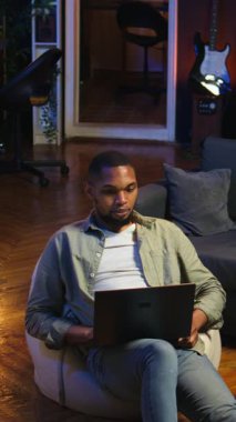 Dikey Video Genç siyah kişi dizüstü bilgisayarında uzaktan görüntülü arama yapıyor evde oturuyor, görevleri çözmek için online konferans webinarına katılıyor. Oturma odasında telekonferans. Kamera.