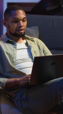 Dikey Video Yetenekli Afro-Amerikan adam havalı oturma odasında dizüstü bilgisayarda çoklu görev yapıyor, serbest çalışma ve uzak sorumlulukları yönetirken internete bağlanıyor. Fasulye torbasında ağ taraması