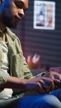 Dikey Görüntü Rahatlatıcı siyah adam rahat bir yerde akıllı telefonuyla kahve masasında oturuyor. Modern ev, şık bir dairede boş zamanların ve çevrimiçi bağlantının altını çiziyor. Kamera.