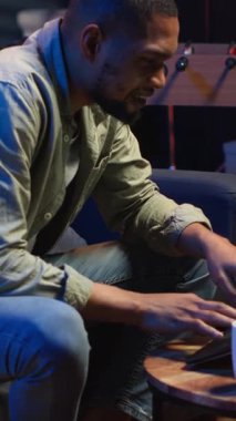 Dikey videolu yetişkin adam dizüstü bilgisayarda yazıyor şık bir dairede koltukta yatıyor, serbest çalışmalar yapıyor ve internetten çalışıyor. Siyahi kişi birden fazla görevi verimli bir şekilde yerine getiriyor ve uzak işin esnekliğini kucaklıyor