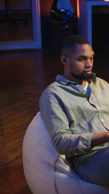 Dikey Video Siyah Adam evde akıllı telefonunu kaydırarak işe ara veriyor, sosyal medyada arkadaşlarına mesaj atıyor ve eğleniyor. Neon ışıkları olan şık bir oturma odasında dinlenen adam. Kamera A.